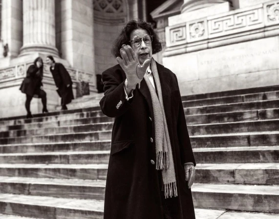 Fran Lebowitz gesturing with her hand in front of the New York Public Library steps, wearing a dark coat and scarf.