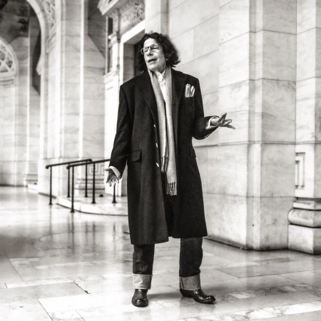 Fran Lebowitz wearing dark overcoat, scarf, and cuffed jeans standing inside a grand classical interior by Michael Avedon