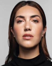 Close-up portrait of a woman with brown hair and glossy lips wearing a black turtleneck against a light grey background.