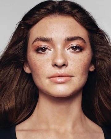 Close-up portrait of a woman with long brown hair, prominent freckles, and light makeup against a neutral background.