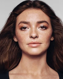 Close-up portrait of a woman with long brown hair, prominent freckles, and light makeup against a neutral background.