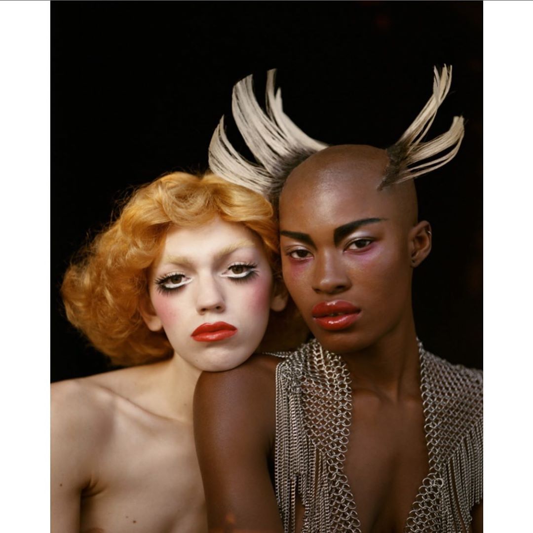 Two models with distinctive makeup and hair pose in a studio, with a blond woman and an African-American woman in the foreground.