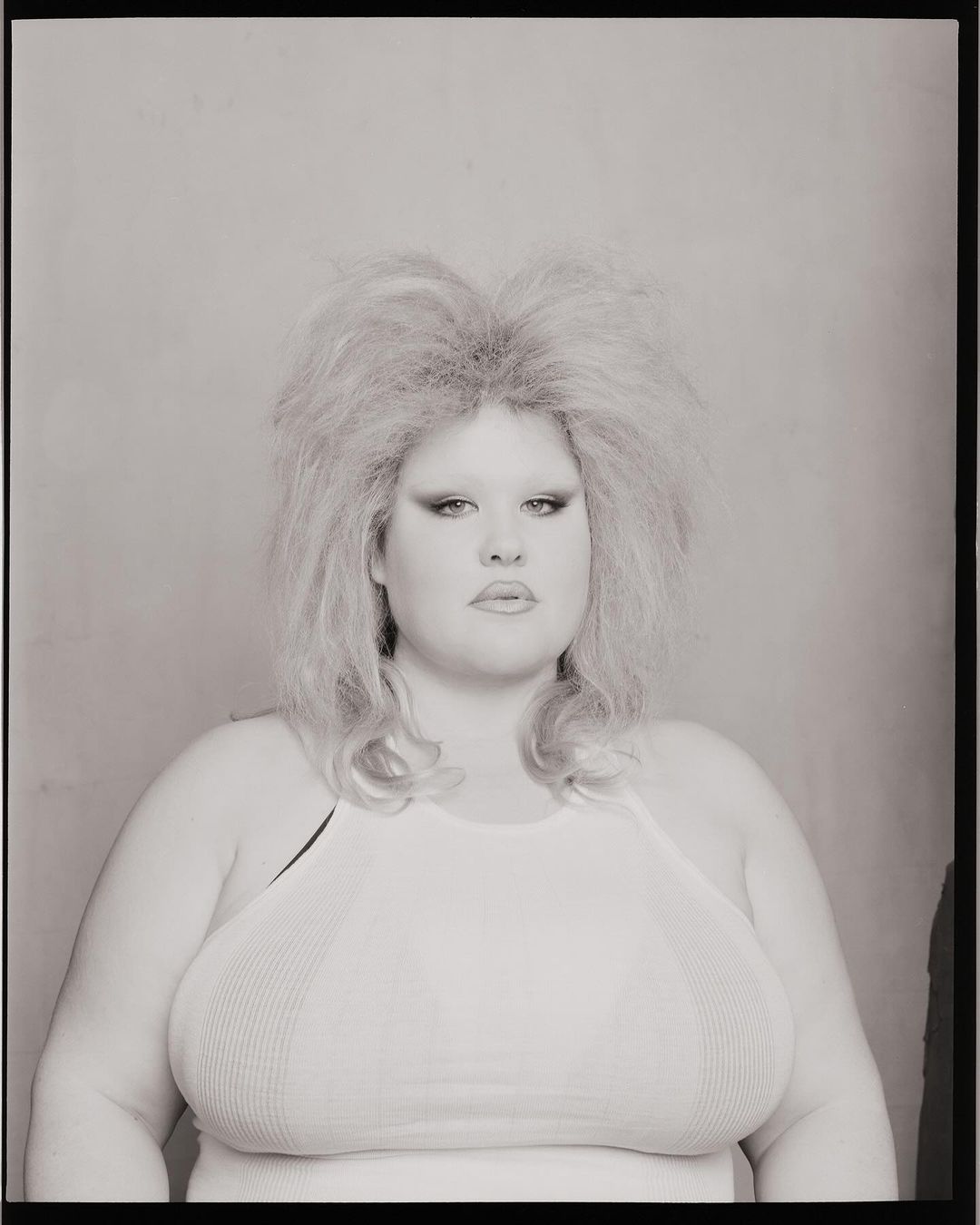 Portrait of a woman with blonde teased hair and a white halter top against a plain background in a black and white photograph.