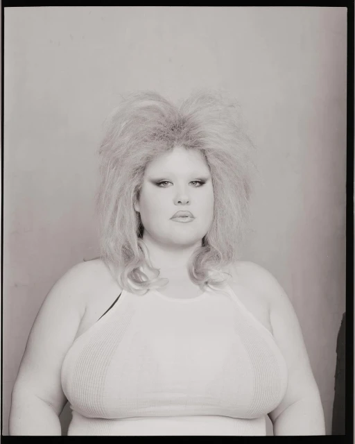 Portrait of a woman with blonde teased hair and a white halter top against a plain background in a black and white photograph.
