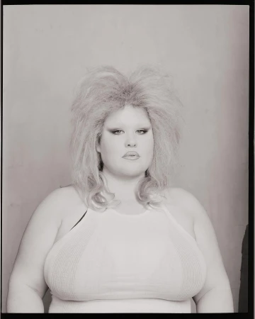 Portrait of a woman with blonde teased hair and a white halter top against a plain background in a black and white photograph.