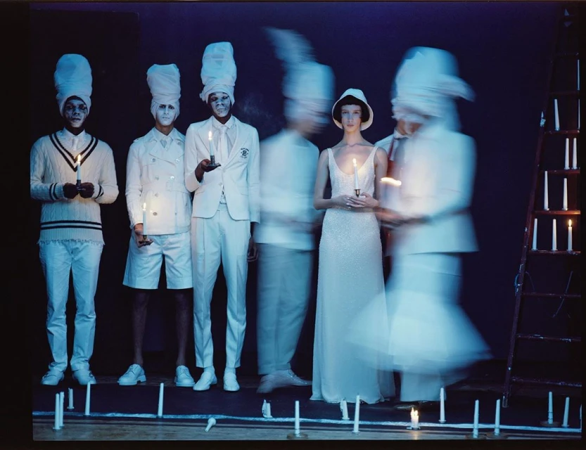 Fashion editorial shot of six models in white outfits, some holding candles, illuminated against a dark backdrop. Ladder with candles.