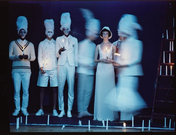 Fashion editorial shot of six models in white outfits, some holding candles, illuminated against a dark backdrop. Ladder with candles.