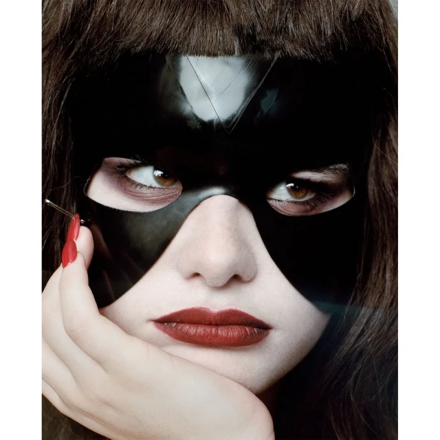 Close-up portrait of a woman wearing a shiny black mask, red lipstick, and dark hair looking directly at the camera.