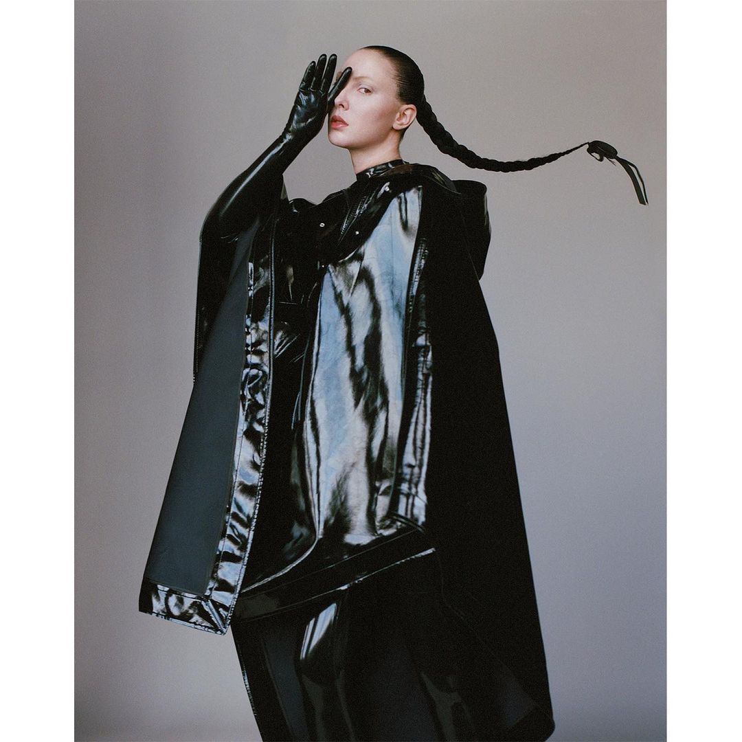 Model in black shiny vinyl cape and gloves with braided hair posing against a neutral grey studio backdrop