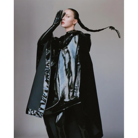 Model in black shiny vinyl cape and gloves with braided hair posing against a neutral grey studio backdrop