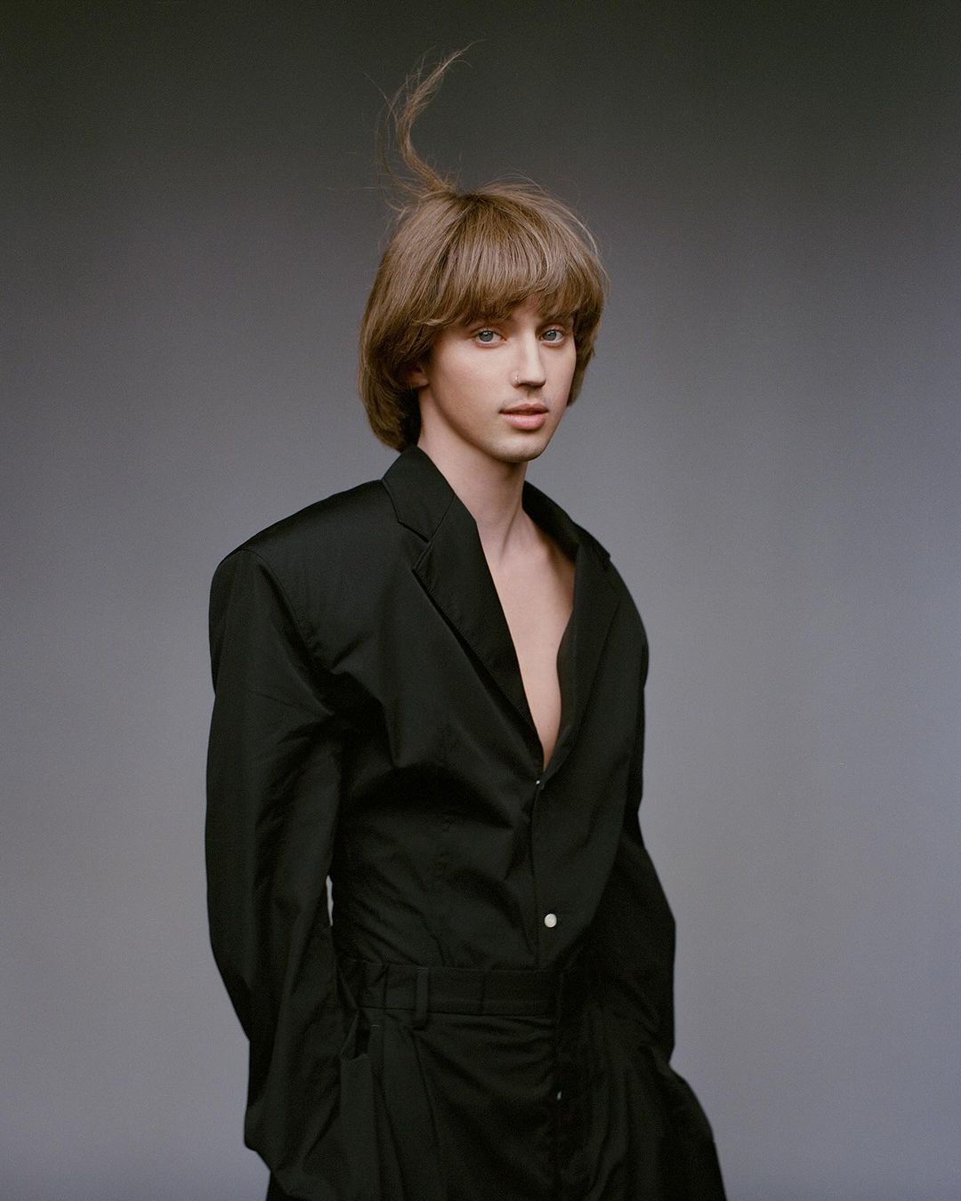 Portrait of a fair-skinned person with brown hair in a black suit, posing in front of a grey studio backdrop with a neutral expression.