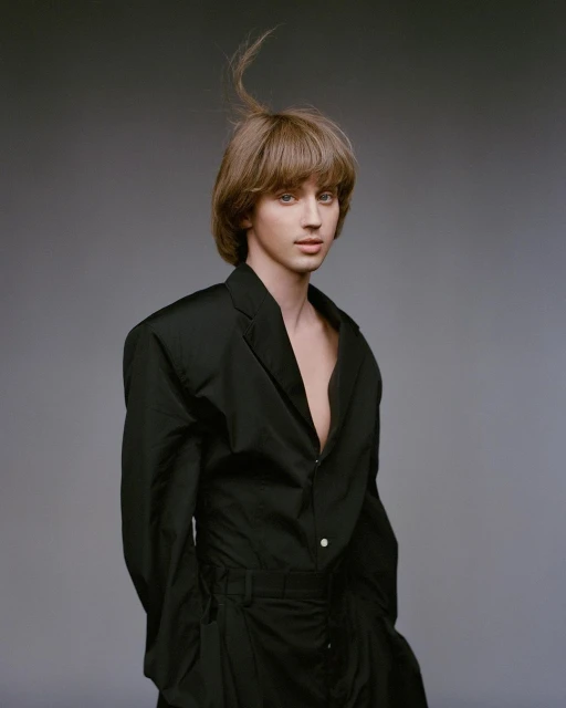 Portrait of a fair-skinned person with brown hair in a black suit, posing in front of a grey studio backdrop with a neutral expression.