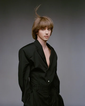 Portrait of a fair-skinned person with brown hair in a black suit, posing in front of a grey studio backdrop with a neutral expression.