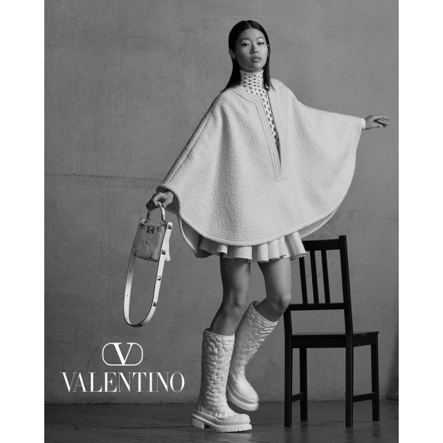 Fashion shot of an Asian woman in a poncho, mini skirt, and boots, holding a purse, posing in front of a monochromatic backdrop.