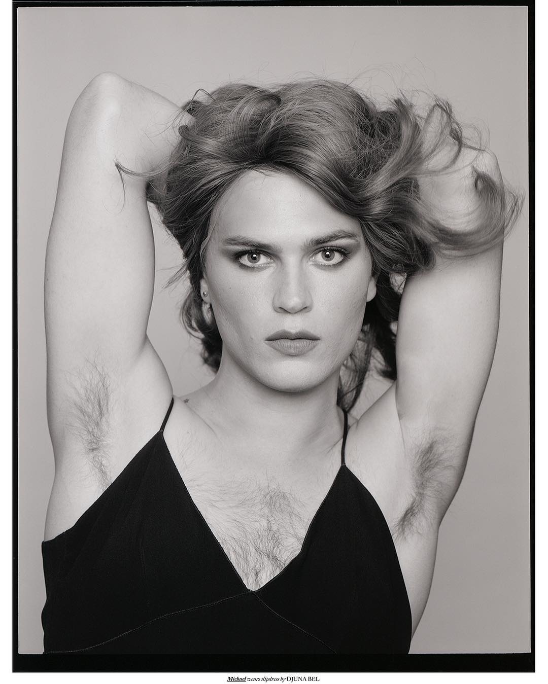 Striking black and white portrait of a young adult with arms raised, set against a neutral backdrop in a studio environment.