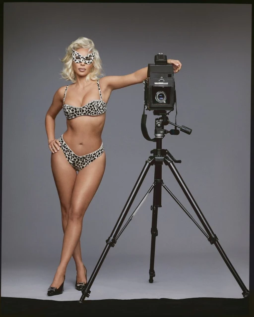 Adult female in leopard print bikini and mask posing with a large format film camera and tripod against a neutral studio background.