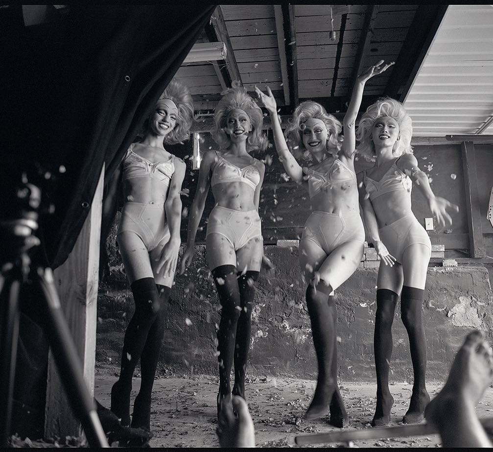 Four women in lingerie and stockings pose in a black and white photograph with feathers falling around them in an indoor environment.