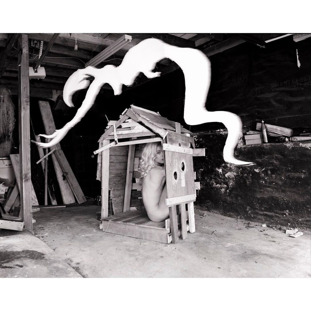 Monochrome photo of a woman inside a wooden structure with an abstract shape hovering above it, set in a cluttered indoor space.