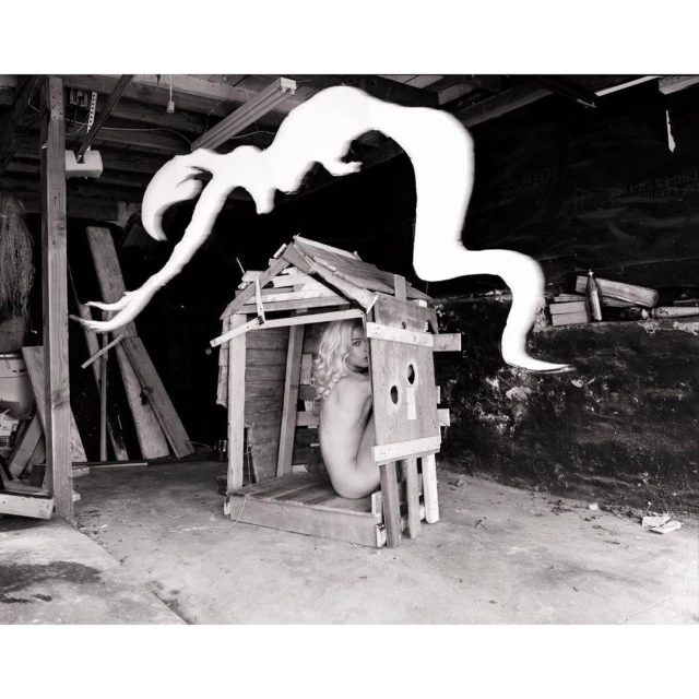 Monochrome photo of a woman inside a wooden structure with an abstract shape hovering above it, set in a cluttered indoor space.