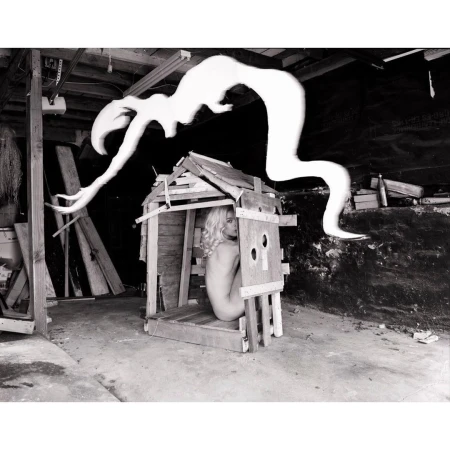 Monochrome photo of a woman inside a wooden structure with an abstract shape hovering above it, set in a cluttered indoor space.