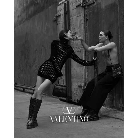 Black and white editorial image of a woman in a mesh dress and boots leaning near a seated man outdoors by a concrete wall.