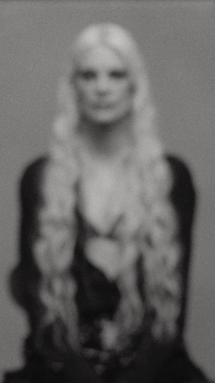 Portrait of a person with long blonde hair and dark clothing captured with heavy soft-focus blur against a plain grey background.