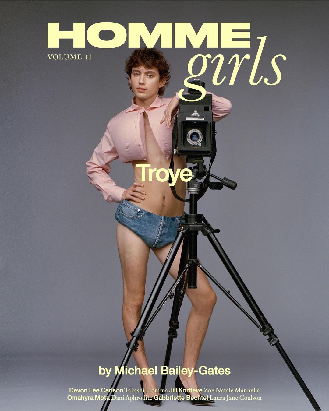 Troye Sivan poses with a vintage camera on a tripod in a studio. He wears a short pink shirt and denim underwear for fashion editorial.