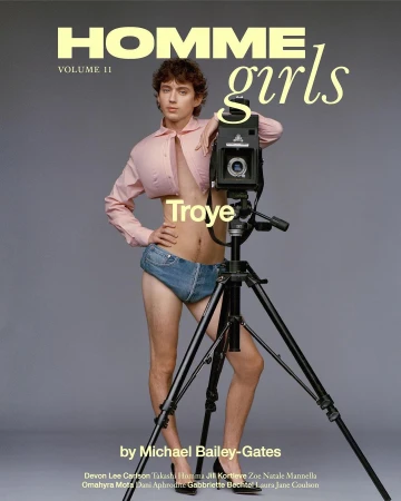Troye Sivan poses with a vintage camera on a tripod in a studio. He wears a short pink shirt and denim underwear for fashion editorial.