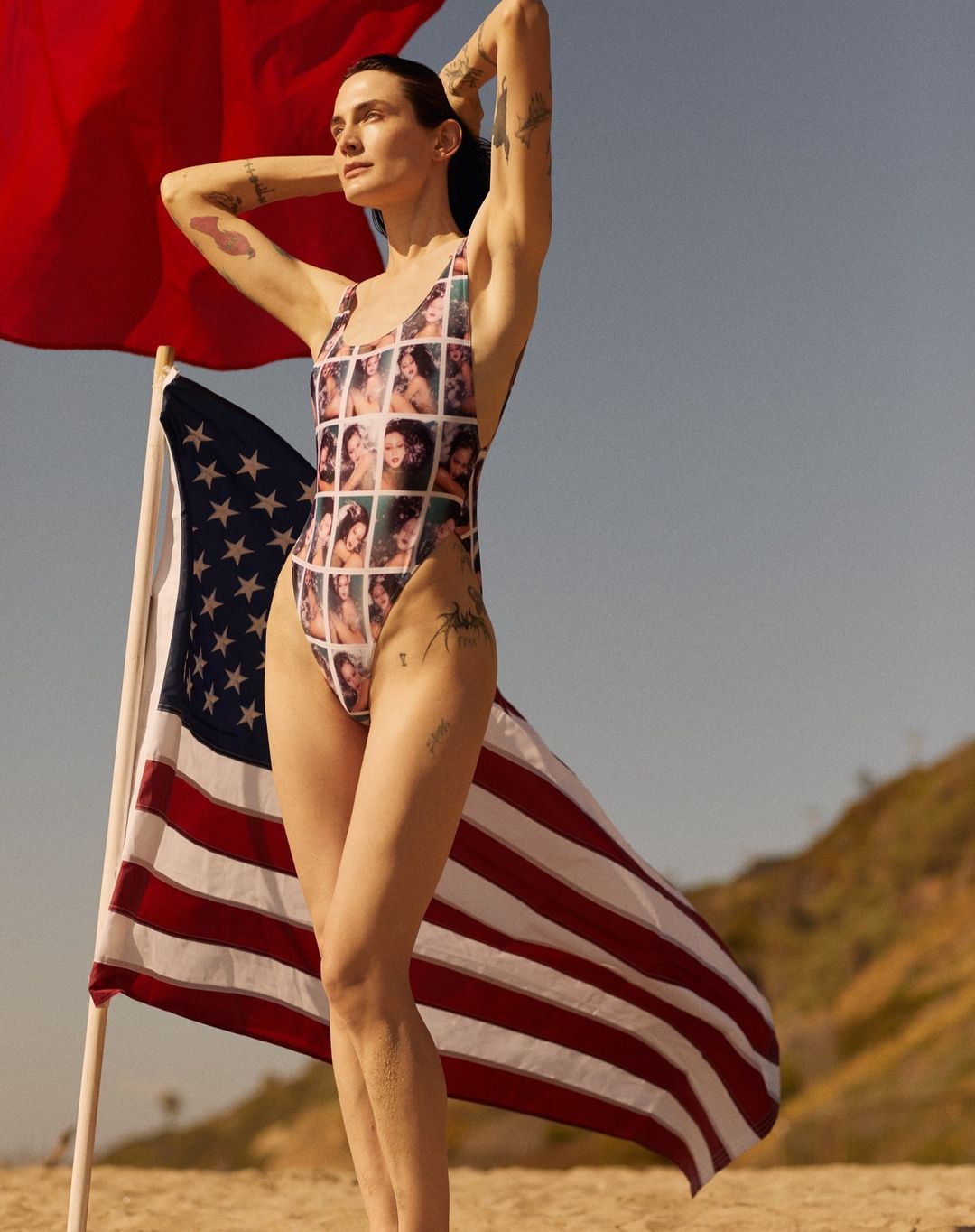 Adult female in swimsuit holding American flag on beach. Woman with arms above head poses in patterned one-piece swimsuit, clear sky.