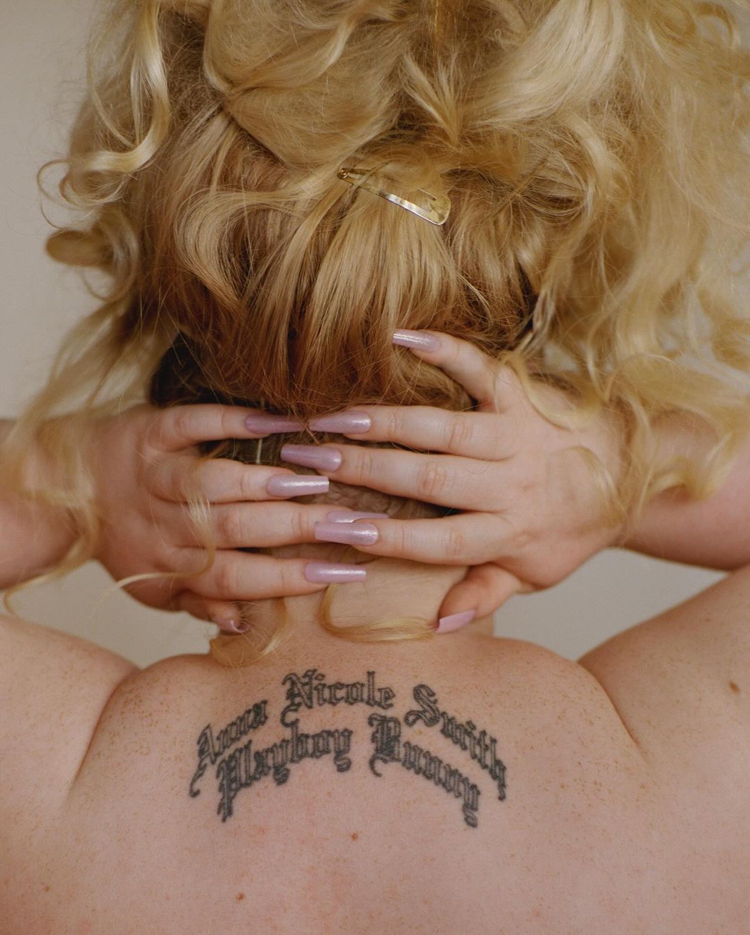 Back view of a woman with blonde curly hair and long pink nails, showing a back tattoo with script text against a neutral background.