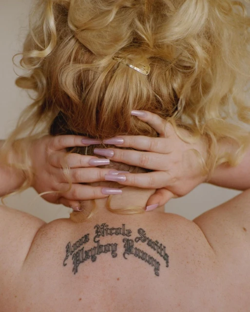 Back view of a woman with blonde curly hair and long pink nails, showing a back tattoo with script text against a neutral background.