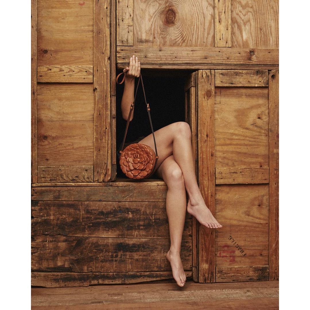Woman's bare legs extend from rustic wood-paneled opening holding a round brown leather purse with floral design details.
