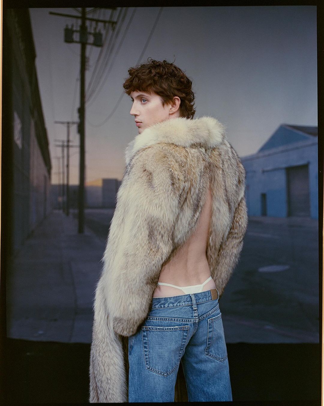 A young adult poses with a fur coat and jeans in a studio setting, looking over their shoulder with a backdrop of a city street.
