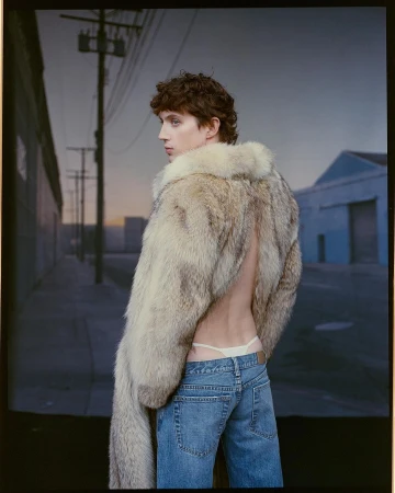 A young adult poses with a fur coat and jeans in a studio setting, looking over their shoulder with a backdrop of a city street.