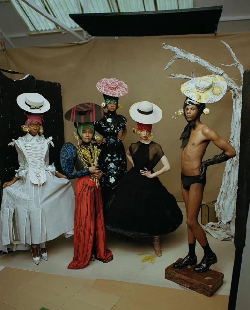 Group of five diverse models in elaborate hats and stylish formalwear striking poses against a neutral backdrop in a fashion photography studio.