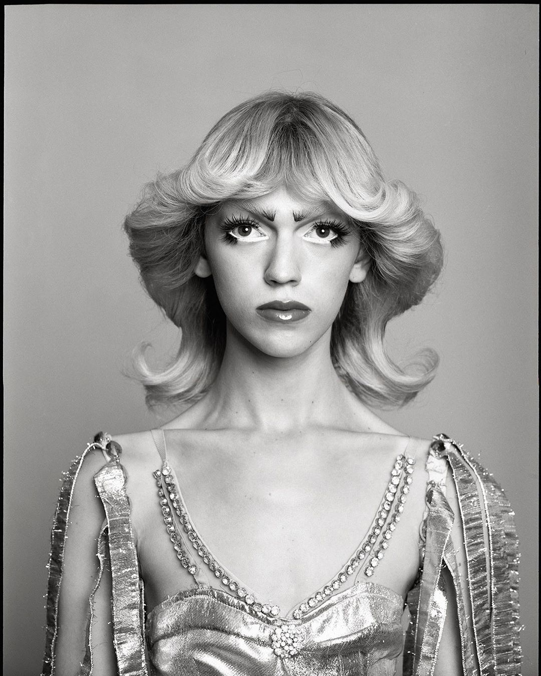 Striking black-and-white portrait of a person with platinum blonde hair, dramatic makeup, and a jeweled dress against a plain background.