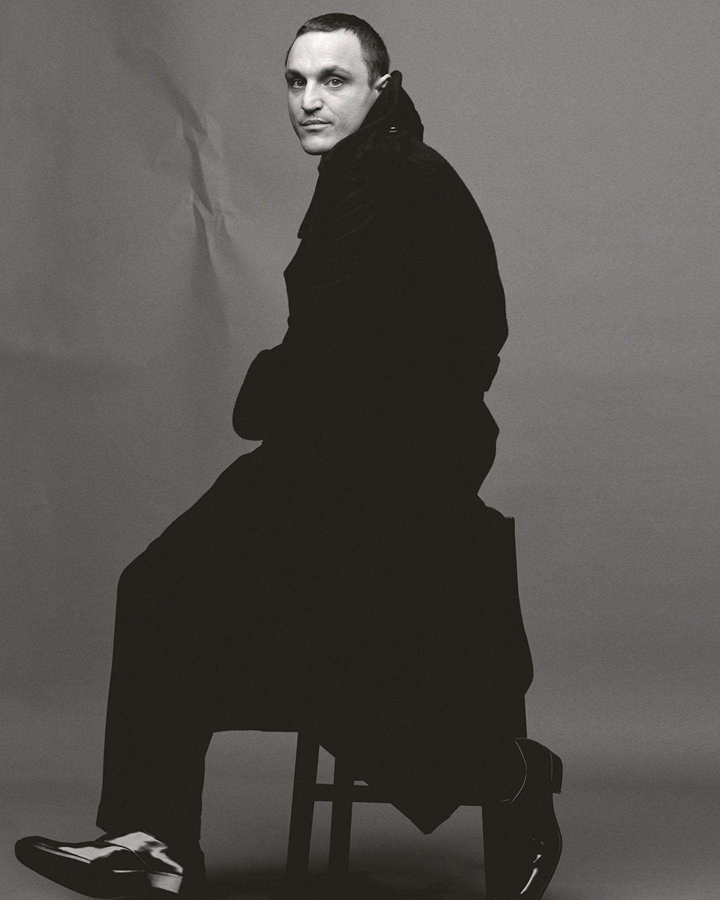 Man wearing a dark trench coat sitting on a stool against a grey seamless paper background in a studio portrait.