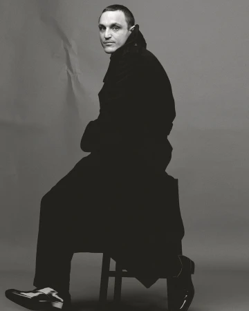 Man wearing a dark trench coat sitting on a stool against a grey seamless paper background in a studio portrait.