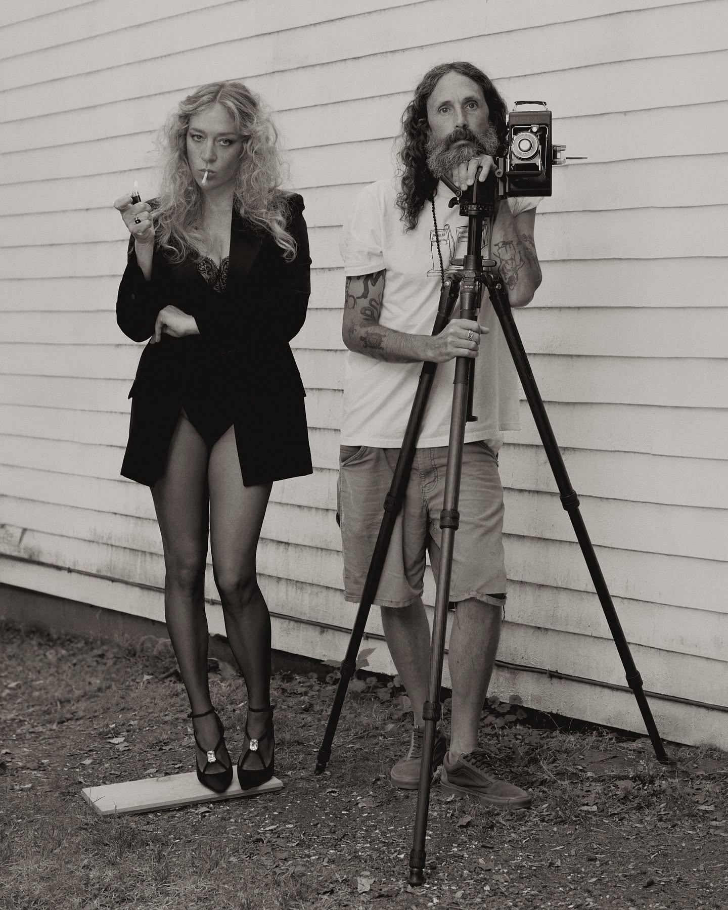A blond woman in a suit lights a cigarette next to a bearded photographer with a vintage camera and tripod against a white wall.