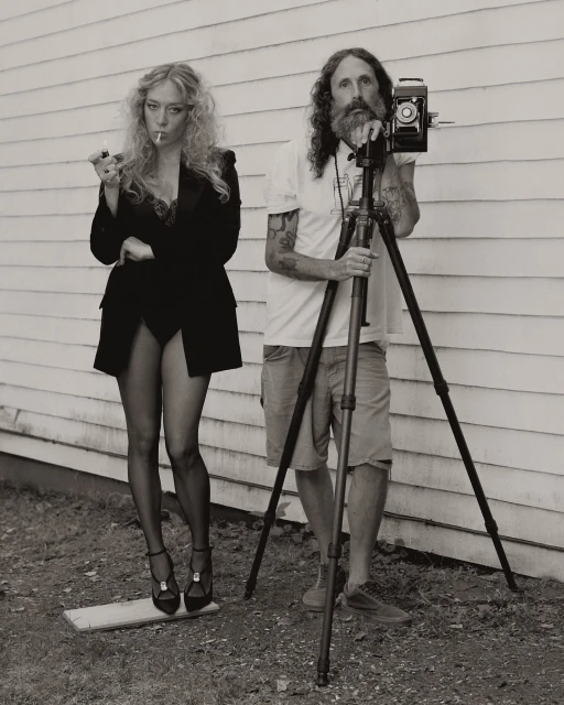 A blond woman in a suit lights a cigarette next to a bearded photographer with a vintage camera and tripod against a white wall.