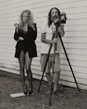 A blond woman in a suit lights a cigarette next to a bearded photographer with a vintage camera and tripod against a white wall.