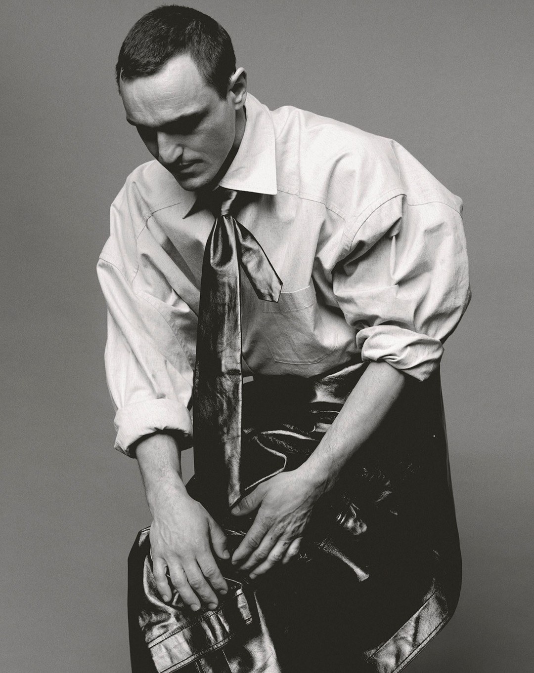 Monochrome portrait of a man wearing a dress shirt and tie, seated against a grey background, looking down with hands clasped.