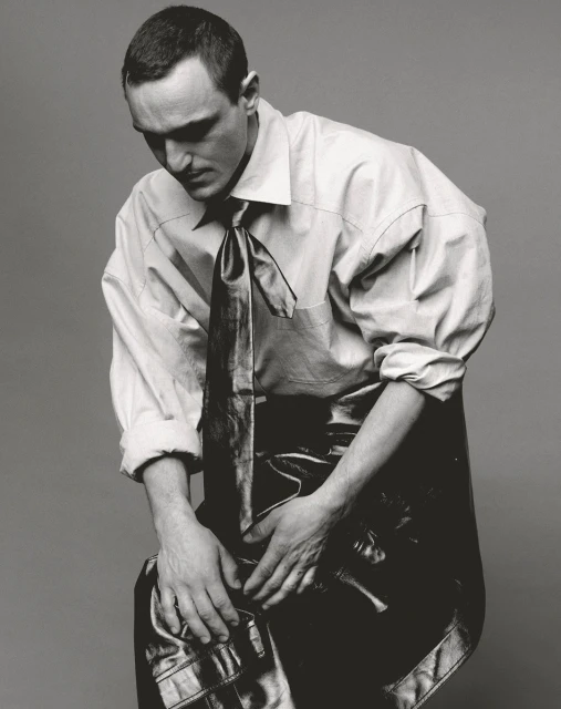Monochrome portrait of a man wearing a dress shirt and tie, seated against a grey background, looking down with hands clasped.