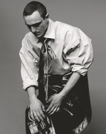 Monochrome portrait of a man wearing a dress shirt and tie, seated against a grey background, looking down with hands clasped.