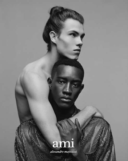 Black and white portrait of two young men in an embrace, one with dark skin wearing a metallic shirt and the other shirtless with fair skin.