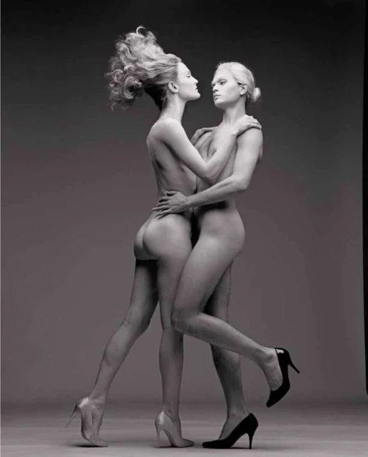 Striking black-and-white studio shot features two nude adult female fashion models with high heels in a unique embracing pose.