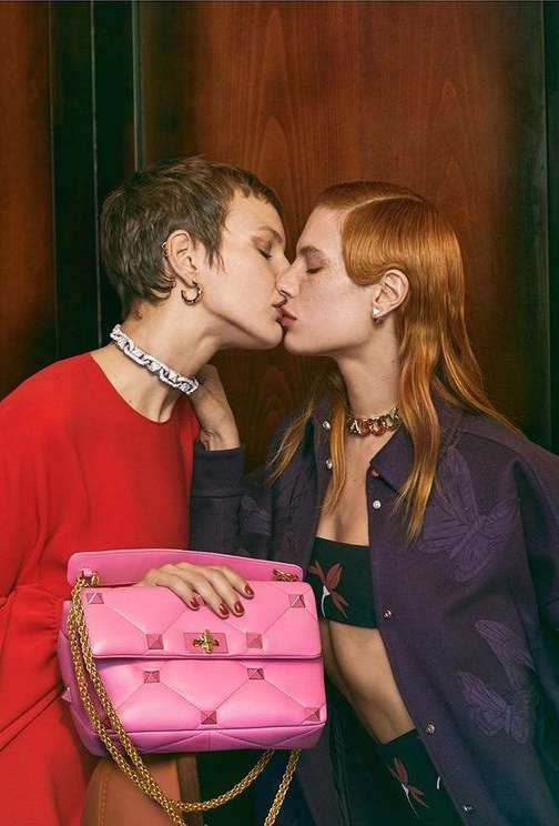 Two adult women kiss while modeling fashion jewelry, pink purse with gold chain, and a dark purple shirt with butterfly pattern.