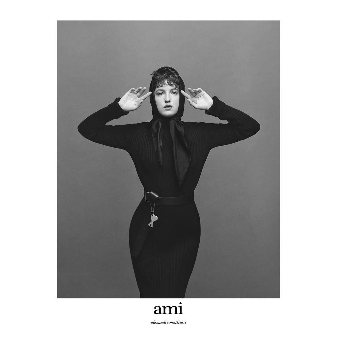 Monochrome portrait of a woman in black dress and headscarf against a gray backdrop, her hands raised to her head in a studio setting.