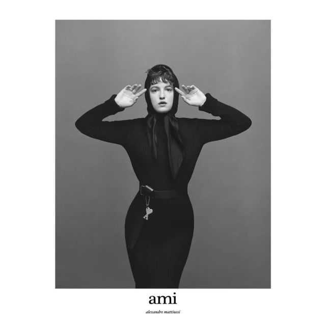 Monochrome portrait of a woman in black dress and headscarf against a gray backdrop, her hands raised to her head in a studio setting.