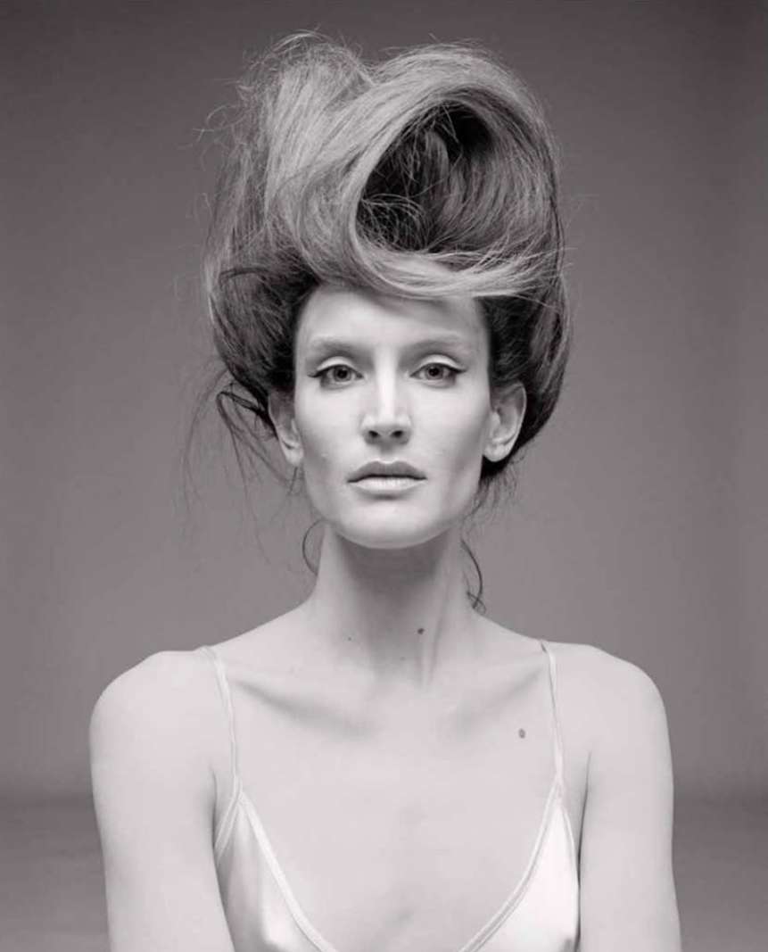 Monochrome portrait of a slender woman with pale skin and an elaborate hairstyle, wearing a satin top, set against a neutral background.